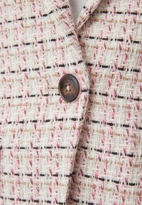 Gros plan sur un blazer en tweed tissé beige, rose et noir avec un bouton marron gravé des mots "REAL IDENTITY."