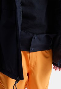 Czarna kurtka o gładkiej fakturze, z zapięciem na zamek błyskawiczny i zatrzaskami. Połączona z pomarańczowymi spodniami, podkreślająca kontrast kolorów.