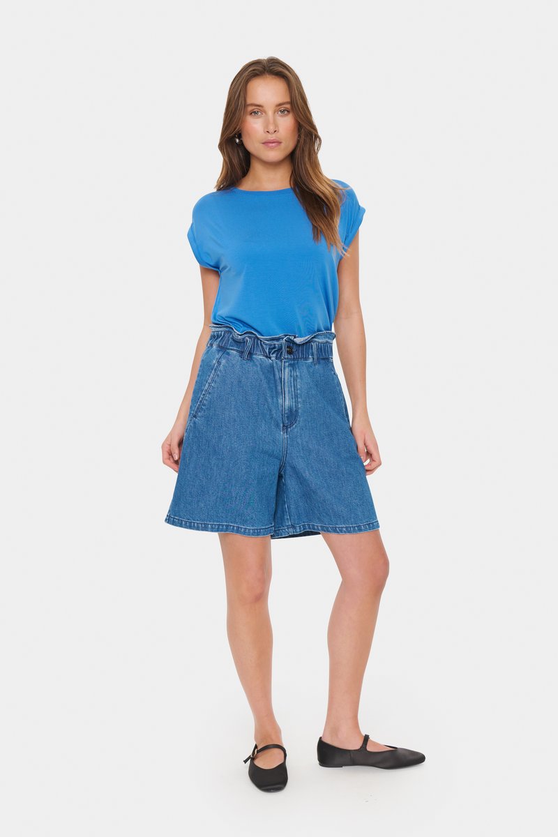 Modra kratka majica s kratkimi rokavi, kombinirana z visokimi denim hlačami v ohlapnem kroju, ki imajo sprednje žepke in gumbasto zapiranje, oblečena s črnimi balerinkami.