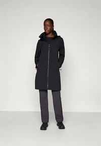 Schwarzer langer Mantel mit hohem Kragen und Frontreißverschluss, ausgestattet mit zwei Seitentaschen und einer glatten, matten Textur. Kombiniert mit dunklen Hosen und Schuhen.