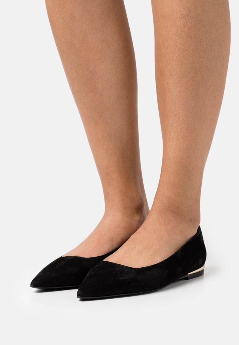 Jambes portant des chaussures plates en daim noir à bout pointu avec un petit détail métallique sur le talon, sur un fond gris clair.