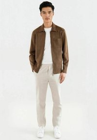 Ung man som står, iförd brun zip-jacka, vit t-shirt, beiga byxor och vita sneakers mot en enfärgad ljus bakgrund.
