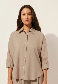Camisa de lino beige de corte holgado con cuello, mangas largas arremangadas, botones frontales con acentos reflectantes de luz y textura suave.
