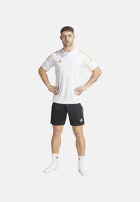 Korte mouw witte sportshirt met gele accenten, gecombineerd met zwarte shorts. Beide items hebben het Adidas-logo. Model staat met handen op de heupen.