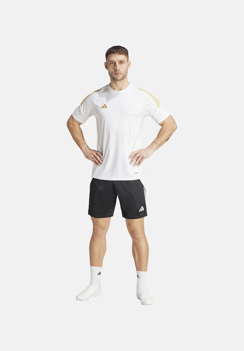 Korte mouw witte sportshirt met gele accenten, gecombineerd met zwarte shorts. Beide items hebben het Adidas-logo. Model staat met handen op de heupen.