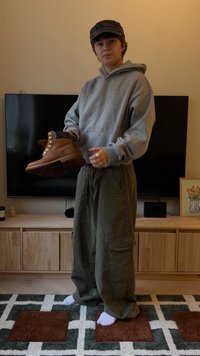 Asmuo, dėvintis pilką džemperį su gobtuvu ir žalias kelnės su kišenėmis, laiko rudą batą gyvenamajame kambaryje, kuriame už jo matomas televizorius ir medinė spintelė.