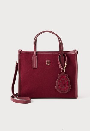 Bolso de mano de fieltro burdeos con doble asa y correa de hombro detachable. Presenta una placa con el logo y un parche con un emblema.