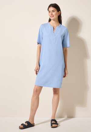 Blau gestreiftes Kurzarmkleid mit einem Reverskragen und seitlichen Schlitzen, aus atmungsaktivem Stoff. Getragen mit dunklen Sandalen.