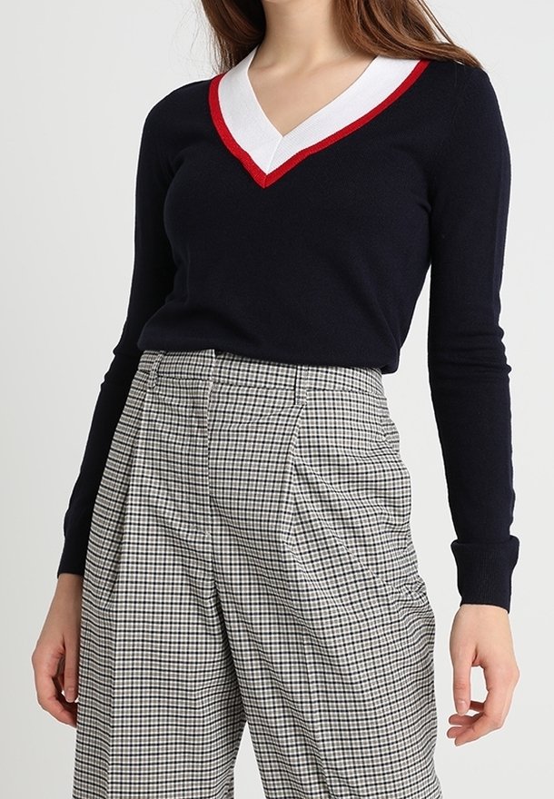 Suéter de punto azul marino con escote en V y trimmings en rojo y blanco, mangas ajustadas, combinado con pantalones de cuadros grises de talle alto con pliegues y piernas anchas.