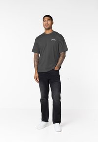 Junger Mann mit tätowierten Armen, der ein dunkelgraues T-Shirt, schwarze Jeans und weiße Sneakers trägt und vor einem einfarbigen weißen Hintergrund steht.