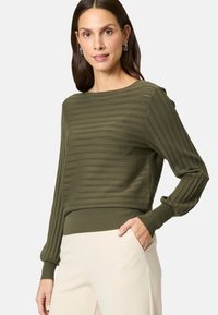 Olijfgroene gebreide trui met een horizontale ribbelstructuur. Heeft lange pofmouwen en een aansluitende tailleband. In combinatie met lichtgekleurde broeken.