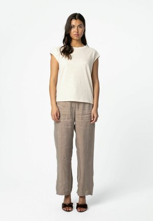 Woman with long dark hair wearing a beige sleeveless top, loose taupe pants, and black open-toe sandals with fur details, standing against white background.