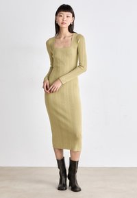 Knit midi dress in olive green with a square neckline, long sleeves, ribbed texture, vertical stripes, and worn with black heeled boots.