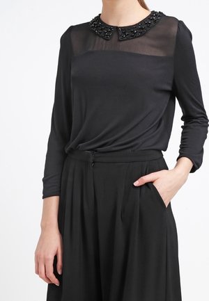 Zwarte blouse met lange mouwen en doorschijnend bovenstuk bij de borst, met een met kralen versierde Peter Pan-kraag, gecombineerd met hoge taille zwarte geplooide broek, met één hand in de zak.