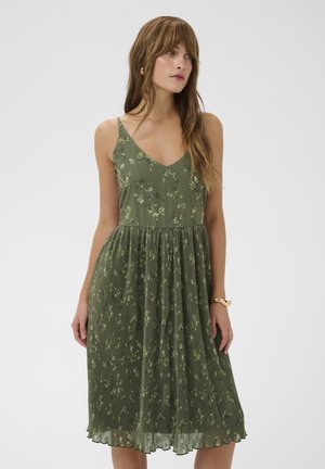 Femme portant une robe midi plissée verte à fleurs avec de fines bretelles, debout devant un fond clair uni, regardant sur le côté.