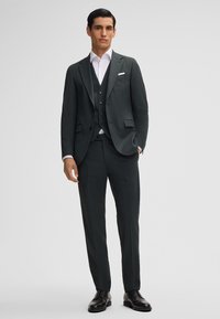 Donkergroen pak met een enkelvoudige blazer, grijze vest en slim-fit pantalon. Een wit shirt en zwarte leren schoenen maken de look compleet.