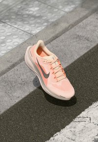 Chaussure de course légère en maille douce pêche avec des accents gris. Elle dispose d'une semelle rembourrée et de lacets plats, conçue pour le confort et la performance.