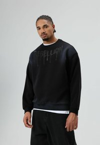 Zwarte sweatshirt met een ronde halslijn, met een zilveren druppelende tekst op de voorkant. Gemaakt van glad, zacht materiaal.