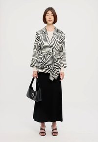 Sort og hvid mønstret blazer med bindebånd i taljen, parret med en ensfarvet sort maxinederdel og sorte hælede sandaler. Modellen holder en sort taske.