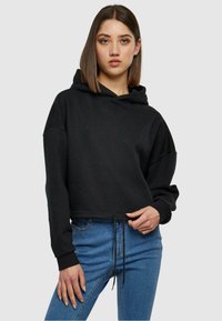 Felpa nera corta con un cordino in vita, dotata di maniche lunghe e realizzata in un tessuto morbido e strutturato. Abbinata a jeans blu.