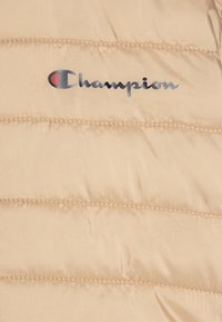 Beige dunjakke med vandrette syninger og en fremtrædende grå "Champion"-logo med farvekontrast. Glat, vandafvisende stof.