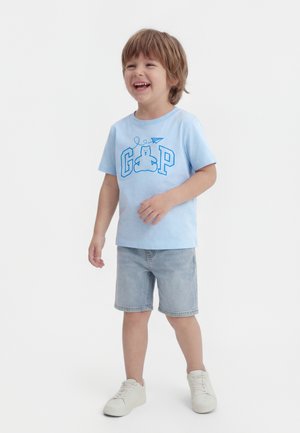 Χαμογελώντας νεαρός αγόρι που φοράει ένα ανοιχτό γαλάζιο μπλουζάκι με εικόνα αρκούδας και χάρτινου αεροπλάνου, τζιν σορτς και λευκά αθλητικά παπούτσια.