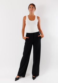 Pantaloni neri a gamba larga con vita alta, dotati di tasche e una texture liscia. Abbinati a un top aderente bianco e tacchi neri.