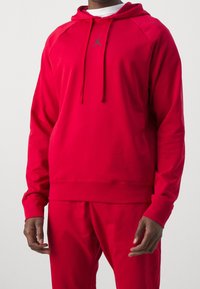 Sweat à capuche rouge en tissu doux, avec une poche kangourou, une capuche à cordon ajustable et un logo discret au centre.