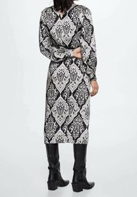 Robe noire et blanche avec un motif damassé, manches longues, décolleté en V et un détail en trou de serrure au dos, accompagnée de bottes noires montantes.