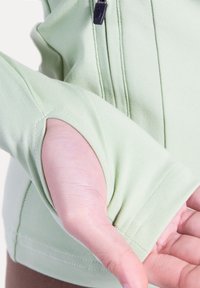 Sudadera de manga larga color verde claro con orificio para el pulgar en el puño. Fabricada con tela suave, presenta detalles sutiles de costura y una cremallera frontal.