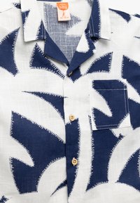 Korte mouwen shirt van witte stof met blauw abstract patroon, voorzien van een zak en houten knopen. De pasvorm is ontspannen met een kraag.