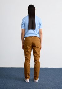 Brązowe spodnie chino o prostym kroju, jasnoniebieska koszulka polo z krótkim rękawem oraz wsuwane buty w kratę, widziane od tyłu.