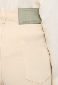 Pantalones beige con un bolsillo trasero y una etiqueta rectangular gris de la marca Trussardi cosida cerca de la cintura.