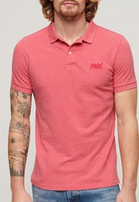Camiseta polo roja hecha de una tela suave, con una tapilla de dos botones y un pequeño logo bordado en el pecho. Mangas cortas con un diseño ajustado.