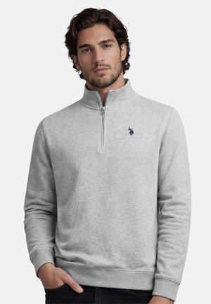 Mann trägt hellgrauen Viertel-Reißverschluss-Pullover mit schwarzem gesticktem Polo-Spieler-Logo, steht vor einfachem weißen Hintergrund.