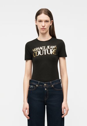 Femeie purtând un tricou negru cu text auriu „VERSACE JEANS COUTURE” și blugi albastru închis, stând în fața unui fundal simplu, deschis.