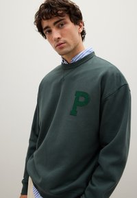 Felpa verde scuro con collo a girocollo, con una lettera "P" ricamata di un verde più scuro. Il tessuto sembra morbido, con una vestibilità rilassata.