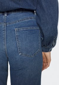 Mörkblå denimjeans med en figurnära design, som har en bakficka och distinkt sömnad. Tyget har en slät yta.