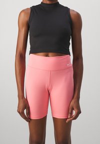 Top corto negro sin mangas con un cuello alto, combinado con pantalones cortos ajustados de cintura alta en rosa claro que presentan un pequeño logo de Nike.