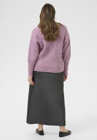 Vrouw met lang bruin haar staat met de rug naar ons gekeerd, terwijl ze een roze gebreide trui, een donkergrijze maxi-rok en zwart platte schoenen draagt, op een witte achtergrond.
