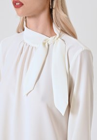 Weiße Bluse mit hohem Kragen und Schleifendetail, aus weichem, glattem Stoff und sanften Falten im Bereich des Kragens.