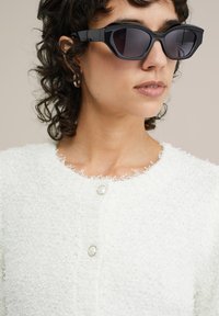 Vit texturerad kofta med en fluffig finish och rundade pärlknappar. Svarta vinklade solglasögon med tonade linser. Lockigt hår synligt.