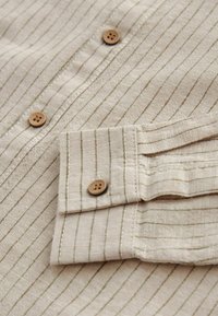Chemise beige à rayures verticales avec des boutons marron clair, montrant les boutons de devant et les détails des poignets sur un tissu texturé.