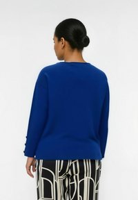 Blauwe trui met een ronde halslijn en lange mouwen, voorzien van knoopdetails bij de mouwen, gecombineerd met gestreepte zwart-witte broeken.