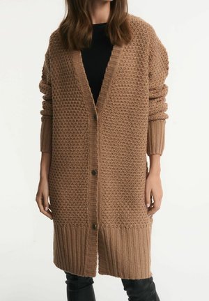 Femme portant un long cardigan en maille épaisse camel avec de gros boutons, poignets et bas côtelés, par-dessus un top noir et un pantalon noir.