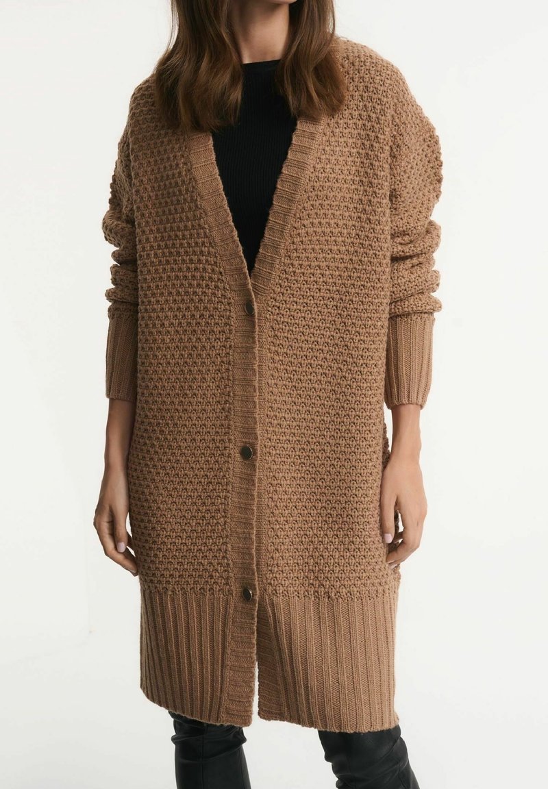 Femme portant un long cardigan en maille épaisse camel avec de gros boutons, poignets et bas côtelés, par-dessus un top noir et un pantalon noir.