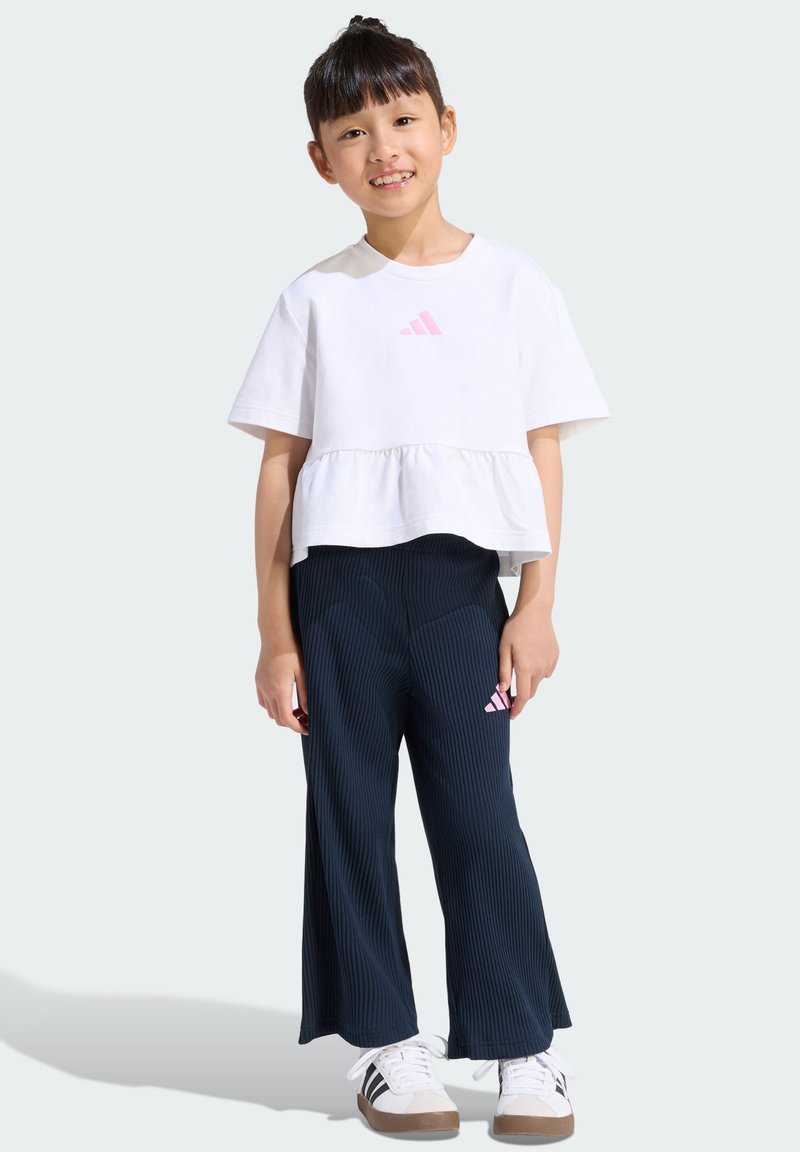 Top blanco de manga corta con logo rosa de Adidas, combinado con pantalones anchos de pierna acanalada en azul marino con logo rosa y zapatillas blancas con suelas marrones.