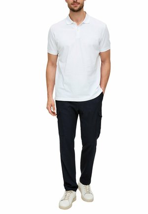 Mann trägt weißes kurzärmeliges Poloshirt, schwarze Cargohosen und weiße Sneakers, steht mit einer Hand in der Tasche.