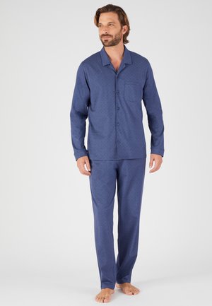Uomo in piedi a piedi nudi che indossa una camicia abbottonata a maniche lunghe e pantaloni coordinati in tessuto blu con un sottile motivo geometrico.