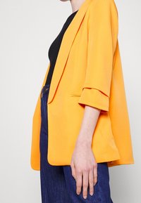Oranžový blazer s uvolněným střihem, mírně oversize tvarem, ohrnutými rukávy a hladkým povrchem. Nositý přes černý top a tmavé džíny.